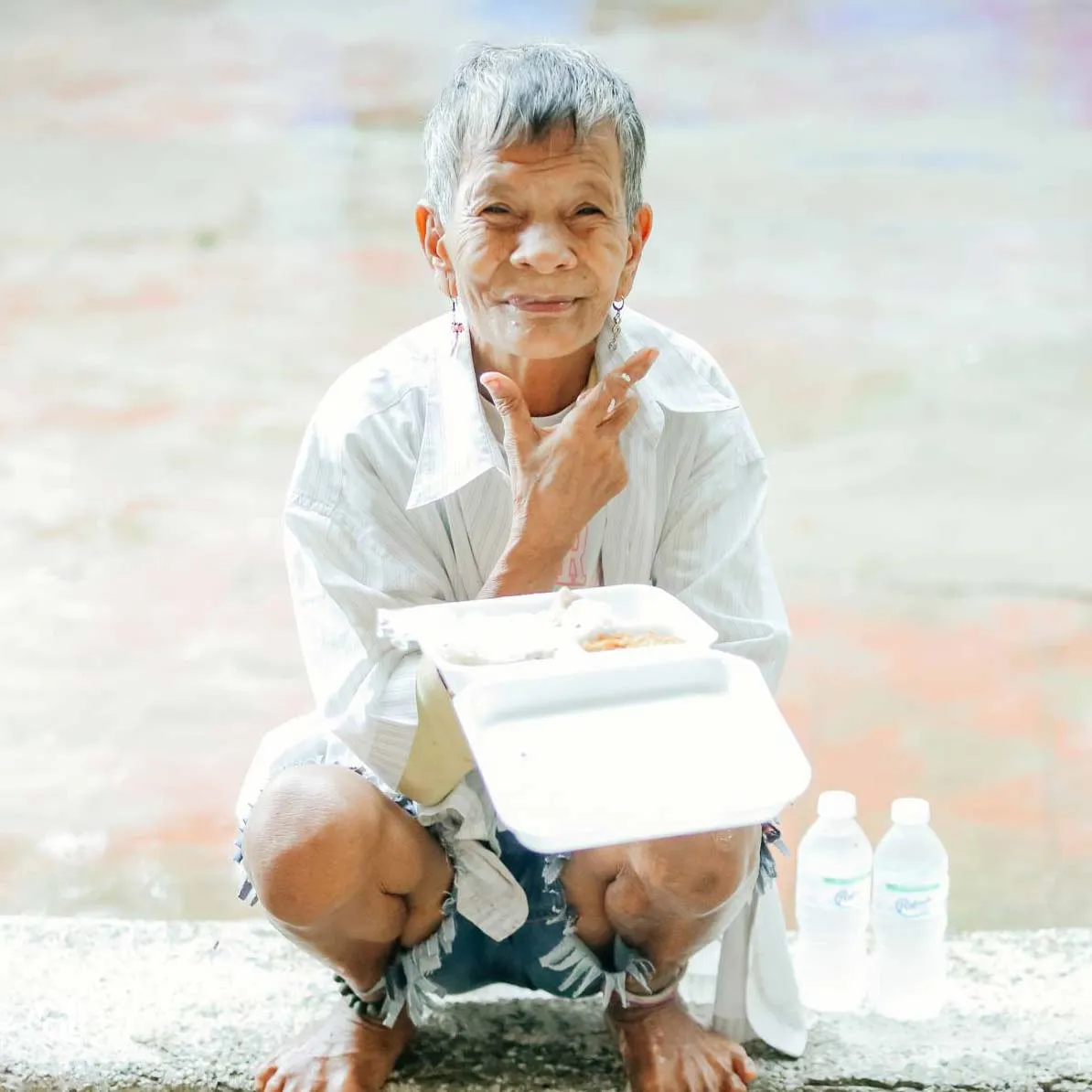 An elderly woman squatting while holding food in a plastic tray, with water bottles next to her.